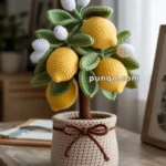 A crocheted lemon tree with yellow lemons and white flowers in a knitted pot on a wooden table.