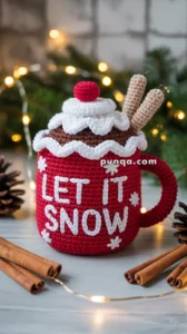 A crocheted red mug with the text 'LET IT SNOW' in white, decorated with a white trim, a red pom-pom, and two beige sticks, set on a festive background with pinecones and string lights.
