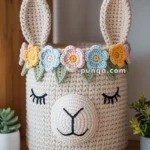 A knitted storage basket in the shape of a cartoon llama's face with closed eyes, adorned with a colorful crocheted flower crown on a wooden shelf.