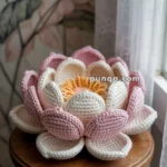 A crocheted lotus flower with pink and white petals and a yellow center, placed on a round wooden stool in front of a floral wallpaper backdrop.