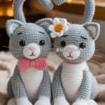 Two crocheted gray and white cats sitting side by side on a beige blanket; one wears a pink bow tie and the other has a white flower with a yellow center by its ear.