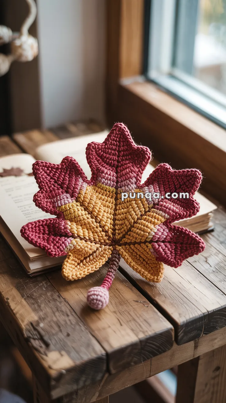 A crocheted maple leaf with red, yellow, and orange hues lies on a wooden table next to an open book by a window.