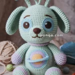 A crocheted plush toy resembling a dog with pastel colors, featuring blue and purple antennas, large round ears, and a colorful Saturn-like planet design on its belly. It is surrounded by yarn balls and small crocheted starfish on a textured surface.