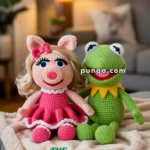 Crocheted dolls of a pig and a frog sitting on a blanket with "The Muppet Show" embroidered on it, set in a warm, cozy room with a lamp in the background.