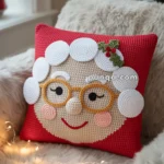 A decorative cushion featuring a crochet design of a smiling face with round glasses and white hair, adorned with holly leaves and berries, set on a red background, placed on a fluffy chair with holiday lights and a Christmas tree in the background.
