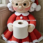 A crocheted Mrs. Claus doll wearing red and white attire, holding a roll of toilet paper, sitting on a tiled bathroom ledge.