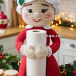 A crochet figure designed like a cheerful, elderly woman holding a roll of paper towels. The figure is dressed in a red and white outfit with Christmas-themed decorations, including a holly accent on her hair. The setup is on a kitchen counter with festive decor in the background.