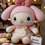 A cute crocheted toy resembling a pink bunny with big black eyes and rosy cheeks sits on a wooden table next to a sign that reads 'Sweet Dreams.' Brightly colored candies are scattered around the toy.