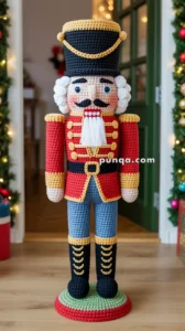 A crocheted nutcracker soldier with a red jacket, blue pants, and a tall black hat stands on a circular base, surrounded by Christmas decorations.