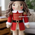 Crocheted toy soldier with a red uniform, tall hat, and long curly hair, standing upright on a patterned surface.