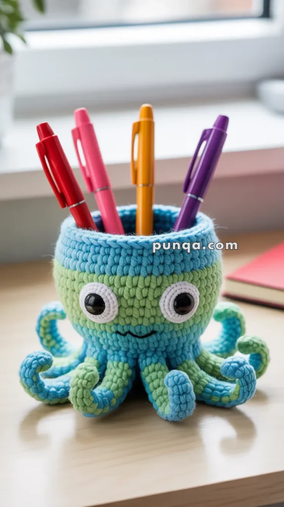 A crocheted octopus-shaped pen holder with large eyes, holding colorful pens on a desk near a window.