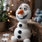 A crochet snowman with an orange carrot nose and stick arms, sitting on a wooden surface next to a sign reading 'let it go' and decorative snowflakes, with a potted plant and string lights in the background.