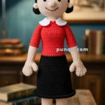 A crocheted doll with a smiling face, wearing a red top with white collar, black skirt, red hat, and yellow shoes, standing on a wooden table with books and a lamp in the background.