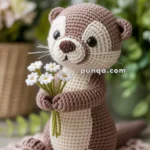 A handmade crochet otter holding a small bouquet of white flowers, sitting on a pink crocheted doily with blurred green and white flowers in the background.