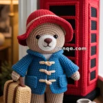 A crocheted teddy bear wearing a red hat and blue coat, holding a beige suitcase, stands next to a crocheted red telephone booth on a table with a small crocheted cup.