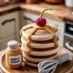 Crocheted stack of pancakes with syrup and a cherry on top, accompanied by a crocheted bottle of maple syrup and a whisk, set on a wooden plate in a kitchen setting.