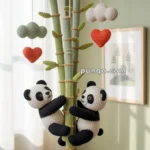 A crochet mobile featuring two pandas climbing a knitted bamboo stalk, along with hanging hearts and clouds, suspended from a circular wooden frame in a softly lit room.