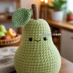 A crocheted pear-shaped plush toy with a smiling face, featuring green yarn for the body and leaf, and brown yarn for the stem, sitting on a wooden table in a cozy kitchen setting.