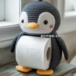 A crocheted penguin figure designed to hold a roll of toilet paper, sitting on a wooden surface by a window.