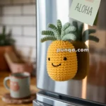 A crocheted pineapple with a smiley face is attached to a refrigerator door, next to a sticky note with the word 'Aloha'. A coffee mug sits on the kitchen counter in the background.