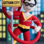 A crocheted figure of a superhero in a red and yellow costume with black eye mask stands next to a crocheted "Gotham City" sign, set against a background of city buildings.
