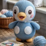 A crocheted blue penguin toy with large eyes and a yellow beak is sitting on a wooden table. Next to it, there is a ball of blue yarn and a book with a Pokémon theme. A wicker basket with yarn balls is in the background.