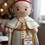 A crocheted doll dressed in papal robes with a red-trimmed hat, holding a sign that reads "God bless you."