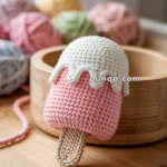 A crochet ice cream-shaped toy with a pink body, white topping, and beige stick, placed on a wooden surface with colorful yarn balls in the background.