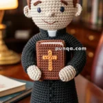 A crocheted doll of a priest with a brown Bible, featuring a cross on the cover, standing on a wooden table in a study room with a desk lamp and bookshelf in the background.