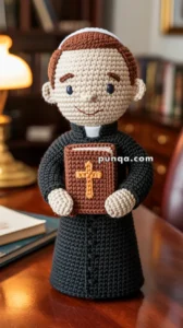 A crocheted doll of a priest with a brown Bible, featuring a cross on the cover, standing on a wooden table in a study room with a desk lamp and bookshelf in the background.