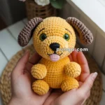 A person holding a small, handmade crocheted dog toy with yellow body and brown ears, featuring green eyes and a pink tongue sticking out, in front of a window with a basket of white and pink roses in the background.