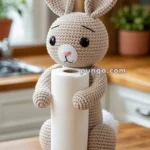 A crocheted bunny holding a roll of paper towels, sitting on a wooden kitchen countertop with a blurred background of kitchen items.