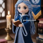 A crocheted doll with long blue hair wearing a blue dress and tiara stands holding a book. It is set in a cozy library environment with books and a candle in the background. A banner is hanging nearby.