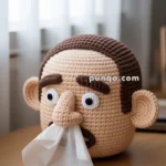 A crocheted tissue box cover shaped like a human head with exaggerated features, including large eyes, ears, and a nose, dispensing tissues from the mouth, placed on a wooden surface in a softly lit room.