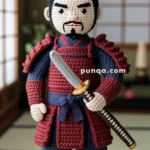 A crochet samurai figurine with a mustache and topknot, wearing red and blue armor and holding a sword, standing in a traditional Japanese room.