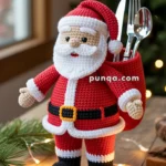 A crocheted Santa Claus figure holding a red sack filled with utensils, placed on a wooden table with Christmas lights and greenery nearby.