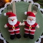 Two crocheted Santa Claus figurines placed on a plate with a green and white crocheted doily, accompanied by silverware. The setting is decorated with pine branches and string lights, creating a festive holiday atmosphere.