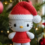 A crocheted Hello Kitty ornament dressed in a red Santa outfit with a white trim, hanging on a Christmas tree branch with festive lights in the background.