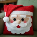 A crocheted pillow in the shape of Santa Claus's face, featuring detailed facial features and a red Santa hat, placed on a green sofa. The background includes a blurred Christmas tree with lights.