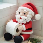 A crocheted Santa Claus doll holding a roll of toilet paper, sitting on a white tiled surface with a small evergreen branch and gold star decoration nearby.