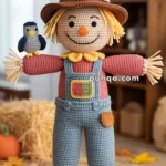 A crocheted scarecrow doll with a cheerful face, wearing a brown hat with a patch, red shirt, and blue overalls stands on a woven mat. A small crocheted bird sits on its arm, and a crocheted pumpkin is on the floor nearby, with autumn leaves in the background.