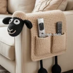 A beige crochet armrest organizer with a black sheep design, featuring pockets holding two TV remote controls, draped over the side of a cream-colored sofa.