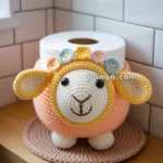 Crocheted sheep toilet paper cover on a bathroom shelf, designed with a cheerful face and floral decorations.