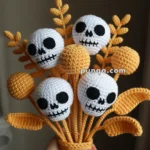 A crocheted bouquet held by a hand, featuring skull faces and yellow decorative elements with leaves and round shapes, against a dark background.