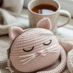 A knitted cat-shaped pouch with closed eyes and whiskers in light pink, resting on a cozy knitted blanket near a cup of tea by the window.