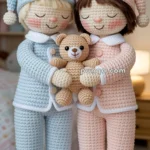 Two crocheted dolls wearing pastel pajamas, one in blue and one in pink, are holding a small crocheted teddy bear between them. They are standing in a cozy bedroom setting with a soft-focus background.