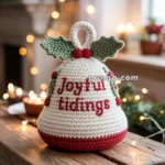 A knitted bell-shaped decoration with "Joyful tidings" written on it, adorned with green leaves and red berries, placed on a wooden table with blurred festive lights in the background.