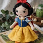 A crocheted doll dressed in a blue and yellow outfit with a red cape and headband, resembling a fairy tale character, standing on an open storybook with blurred plants in the background.