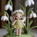 A crocheted fairy doll with wings, wearing a green dress and a floral crown, stands among crocheted snowdrop flowers on a round wooden base.
