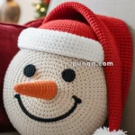 A crocheted snowman pillow with a red Santa hat rests on a beige couch, with holiday decorations blurred in the background.